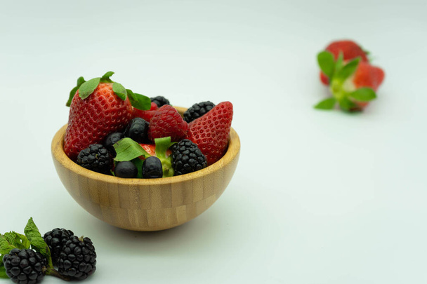 View of wooden bowl full of strawberries, blackberries, raspberries and blueberries with strawberries behind.White background, space for text. Healthy food concept. - Photo, image
