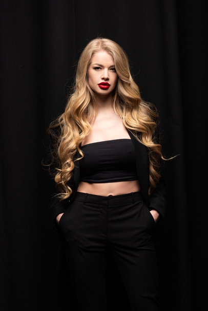 stylish girl with red lips standing with hands in pockets isolated on black  - Фото, зображення