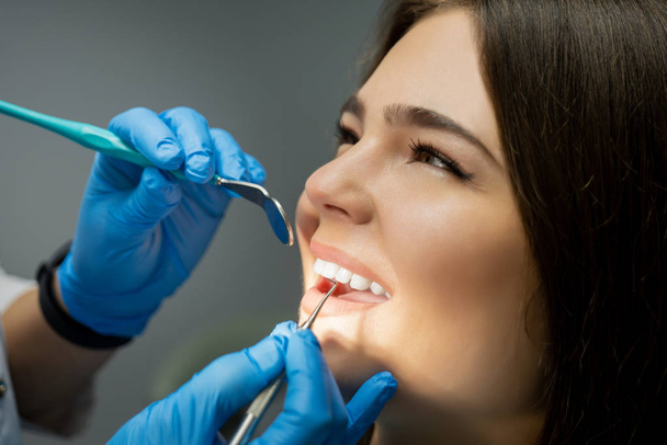 smiling happy brunette woman patient examined by dentist in blue gloves using dental mirror and scaler sitting half a turn in dental chair , healthcare concept - Photo, image