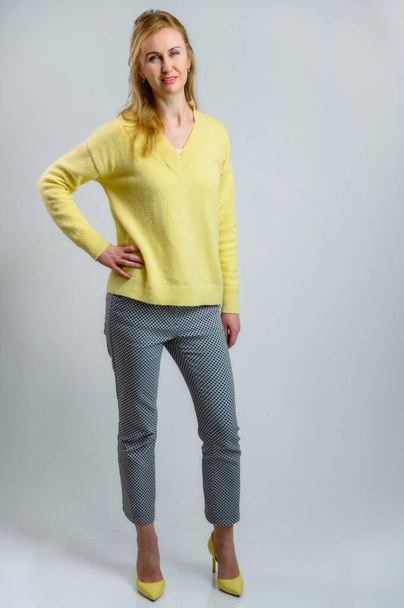 Full-length portrait of a stylish pretty blonde woman in a yellow sweater and gray pants on a white background - Foto, afbeelding