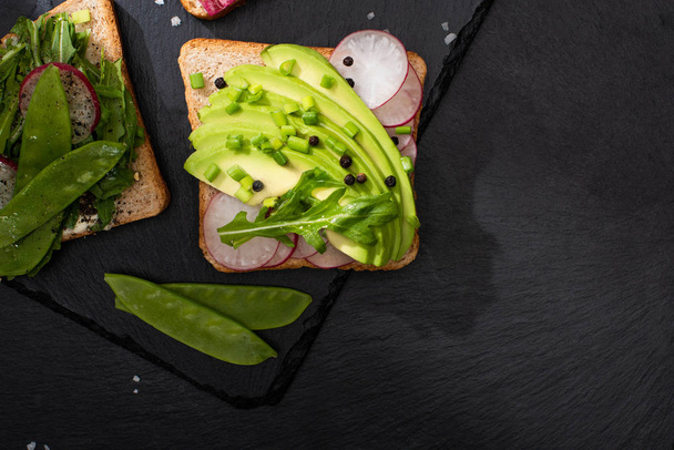 bovenaanzicht van verse broodjes met radijs en avocado op stenen bord met peper en zout - Foto, afbeelding