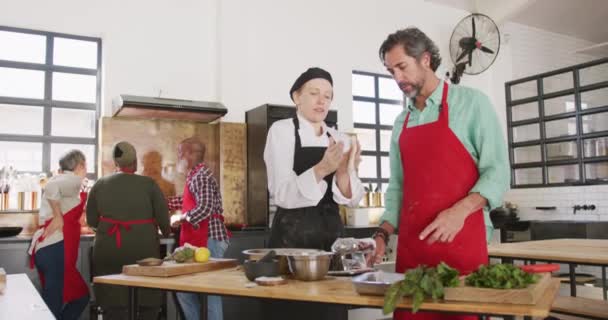 Front view of a senior Caucasian man and a Caucasian female chef during cookery class in a restaurant kitchen, the chef explaining and showing the man how to roll dough through a pasta machine, in slow motion - Imágenes, Vídeo