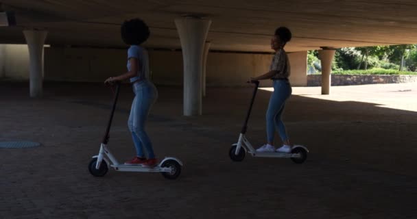 Side view of two mixed race women enjoying free time in a urban park on a sunny day together, riding electric scooters, slow motion - Filmagem, Vídeo