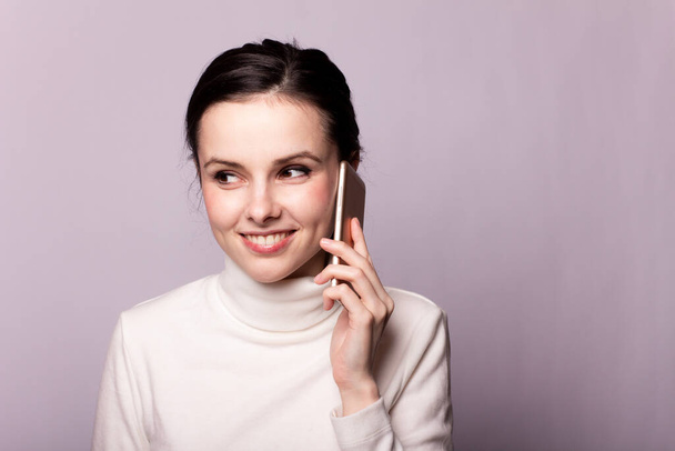 girl in a white turtleneck talking on the phone, portrait on a gray background - Zdjęcie, obraz