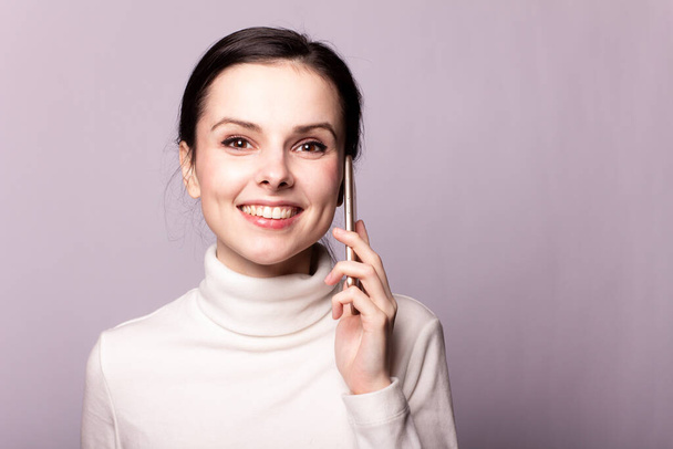 girl in a white turtleneck talking on the phone, portrait on a gray background - Фото, изображение