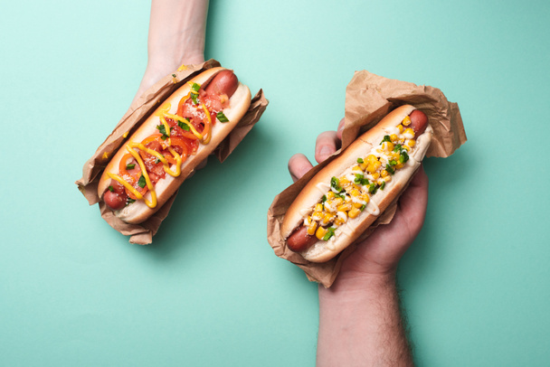 cropped view of man and woman holding two tasty hot dogs in paper on blue - Photo, Image