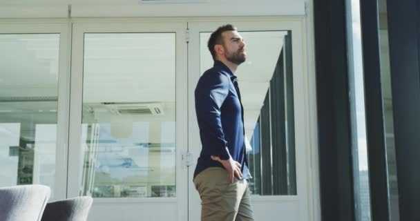 Side view of a Caucasian businessman working in a modern office, taking a break standing by the window with hands on hips admiring the view in slow motion - Imágenes, Vídeo