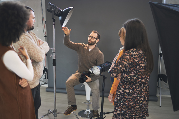 Jeune tuteur barbu attrayant assis sur une chaise en studio, tenant la caméra et expliquant quelque chose sur la photographie pendant que les participants debout et le regardant
. - Photo, image