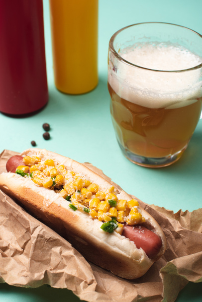 close-up van hotdog met maïs, glas bier en flessen met mosterd en ketchup op blauw - Foto, afbeelding