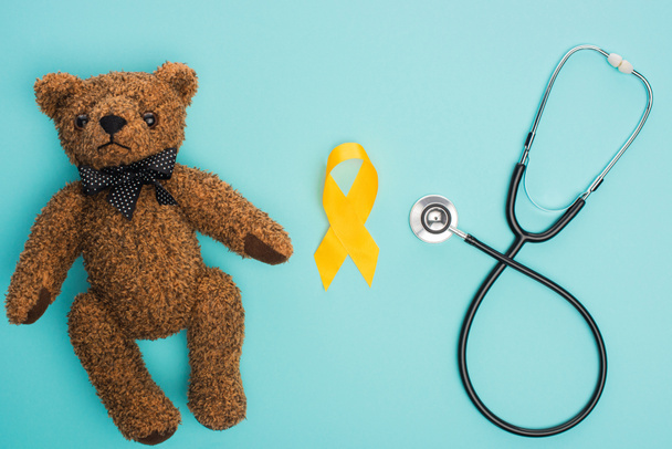 Top view of yellow awareness ribbon, teddy bear near stethoscope on blue background, international childhood cancer day concept - Photo, Image