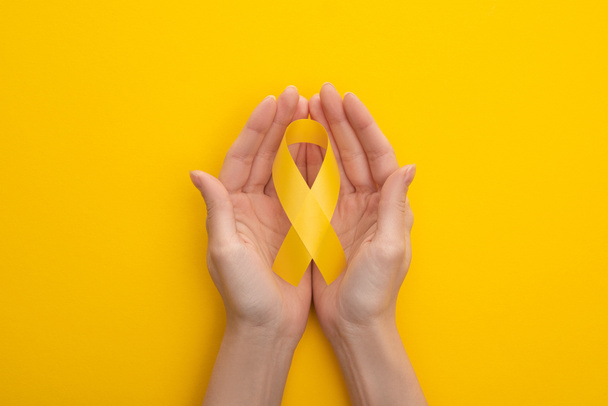 Częściowy widok żeńskich rąk z żółtą wstążką świadomości na kolorowym tle, koncepcja międzynarodowego dnia nowotworu w dzieciństwie - Zdjęcie, obraz