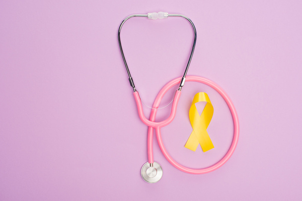 Vista superior de la cinta amarilla con estetoscopio rosa sobre fondo violeta, concepto del día internacional del cáncer infantil
 - Foto, Imagen