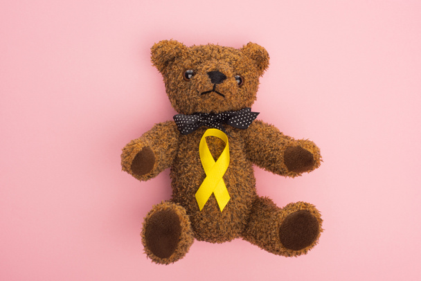 Vista dall'alto del nastro di consapevolezza giallo su orsacchiotto con fiocco su sfondo rosa, concetto di giornata internazionale del cancro infantile
 - Foto, immagini
