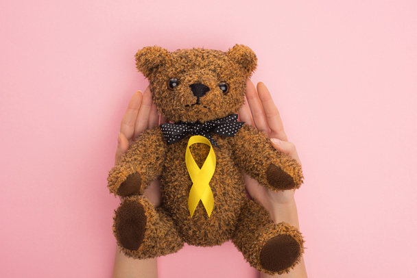 Oříznutý pohled ženy držící plyšového medvídka se žlutou stuhou na růžovém pozadí, mezinárodní koncepce dne rakoviny v dětství - Fotografie, Obrázek