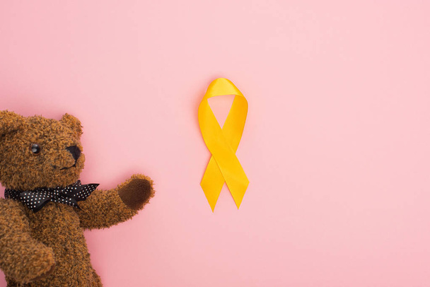 Horní pohled na žlutou stuhu a hračku na růžovém pozadí, mezinárodní koncepce dne rakoviny dětství - Fotografie, Obrázek