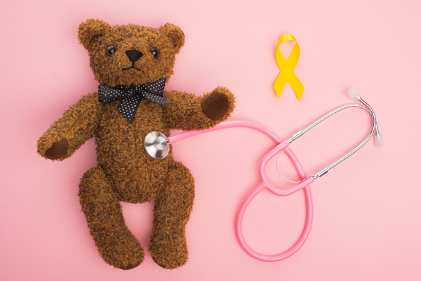 ピンクを背景にテディベアとつながる黄色のリボンと聴診器のトップビュー、国際小児がんの日のコンセプト - 写真・画像