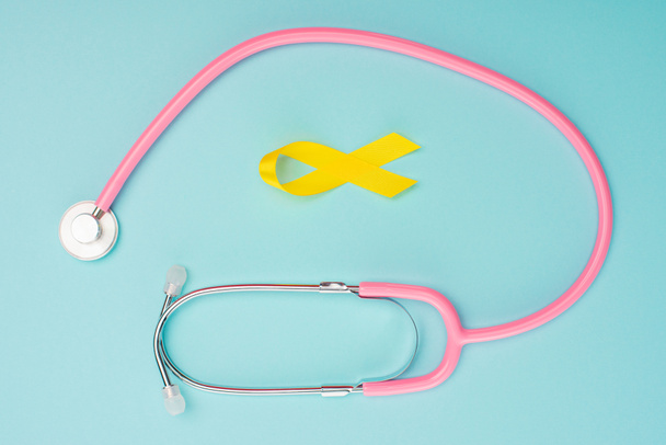 Top view of yellow ribbon and stethoscope on blue background, international childhood cancer day concept - Fotografie, Obrázek