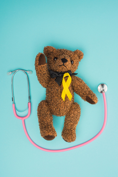 Top view of yellow ribbon on brown teddy bear near stethoscope on blue background, international childhood cancer day concept - Photo, image
