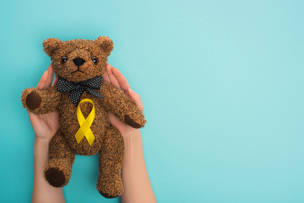 Cropped view of woman holding teddy bear with yellow ribbon on blue background, international childhood cancer day concept - Photo, Image