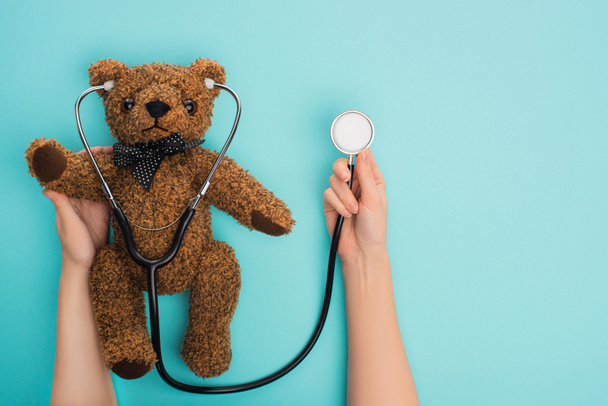Cropped view of woman holding teddy bear with stethoscope on blue background, international childhood cancer day concept - Photo, Image