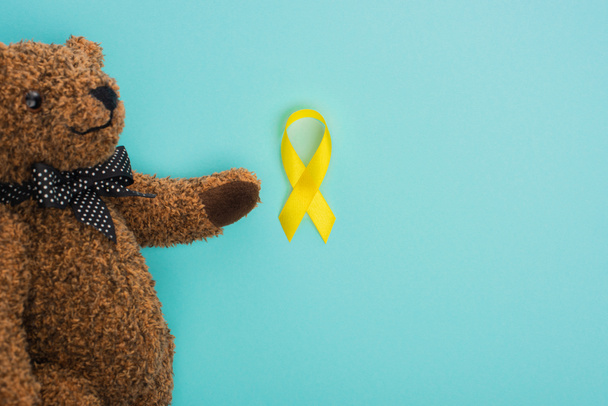 Top view of teddy bear with bow and yellow ribbon on blue background, international childhood cancer day concept - Photo, Image