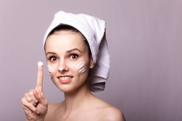 girl with a white towel on her head and cream on her face and hand, gray background - Foto, afbeelding