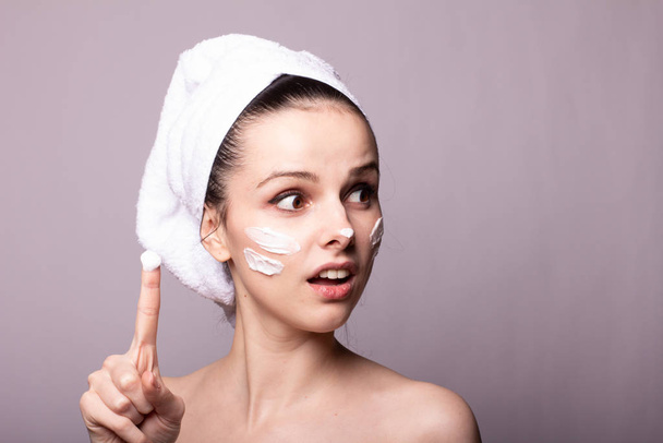 girl with a white towel on her head and cream on her face and hand, gray background - Photo, Image