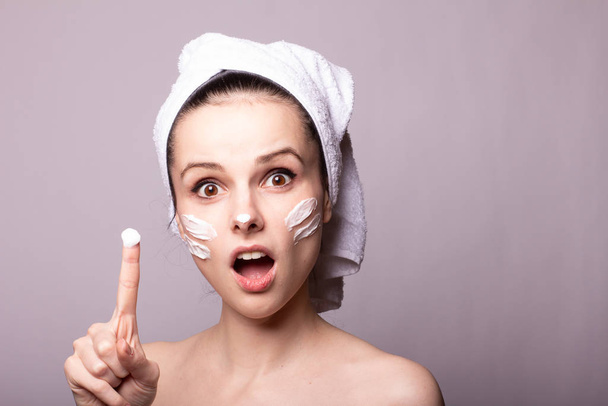 girl with a white towel on her head and cream on her face and hand, gray background - Foto, Bild
