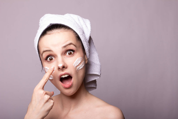 girl with a white towel on her head and cream on her face and hand, gray background - Foto, Imagem