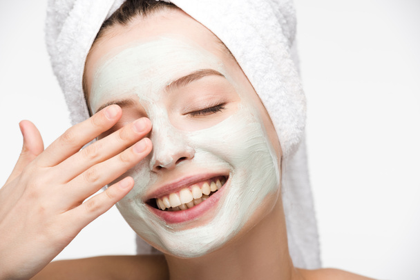 happy girl with facial nourishing mask and towel on head touching eye isolated on white - Photo, Image