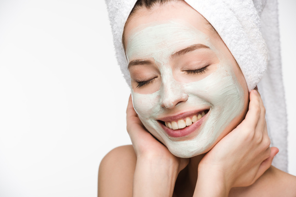 smiling girl with facial nourishing mask and towel on head touching face with closed eyes isolated on white - Фото, изображение