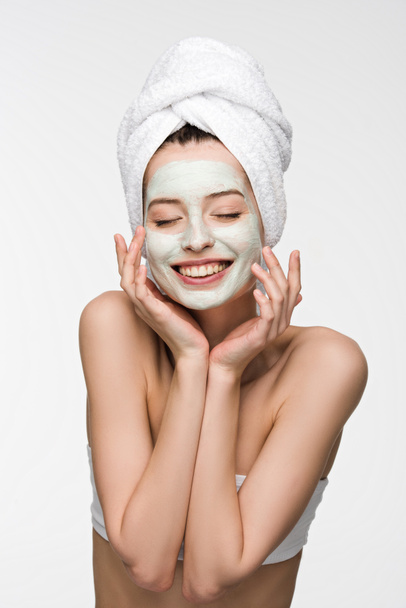 veselá dívka s vyživující obličejovou maskou a ručníkem na hlavě dotýkající se obličeje se zavřenýma očima izolované na bílém - Fotografie, Obrázek