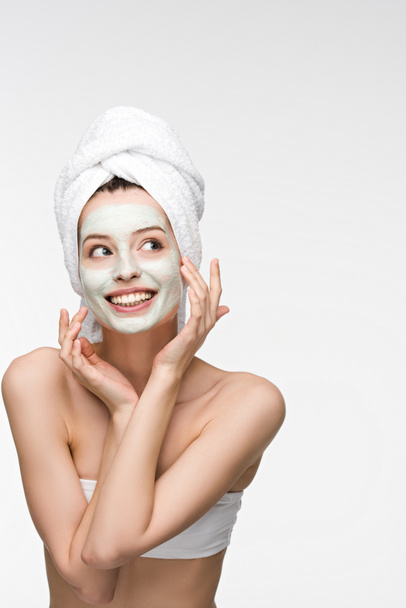 smiling girl with nourishing facial mask and towel on head touching face isolated on white - Photo, Image