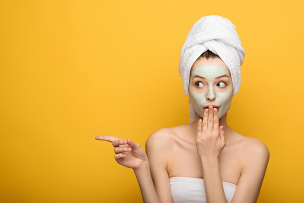 shocked girl with nourishing facial mask and towel on head covering mouth with hand and pointing with finger on yellow background - Foto, afbeelding