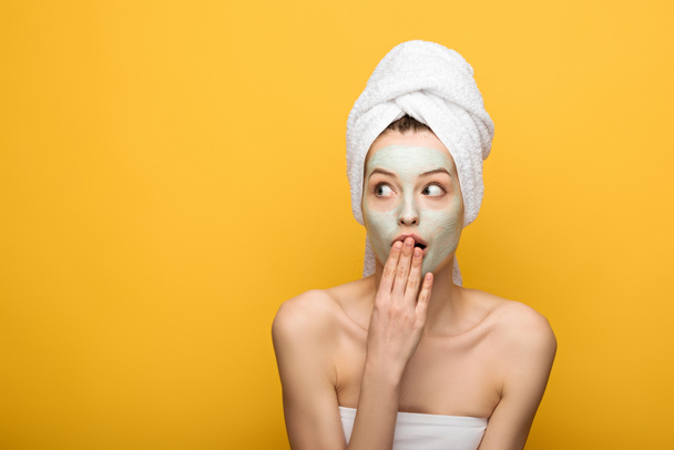 shocked girl with nourishing facial mask covering mouth with hand and looking away isolated on yellow - Photo, Image