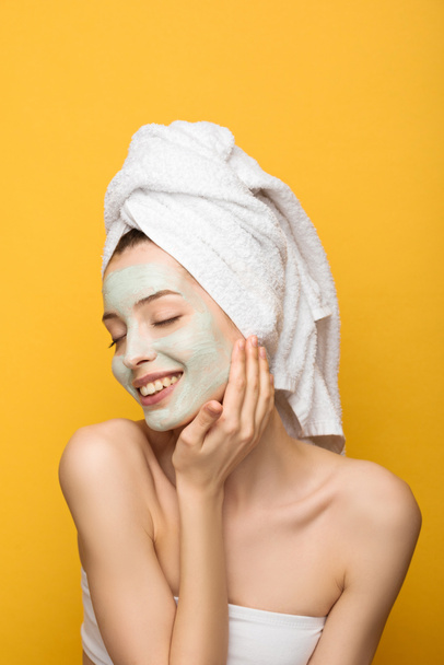 happy girl with nourishing facial mask and towel on head touching head with closed eyes isolated on yellow - Φωτογραφία, εικόνα
