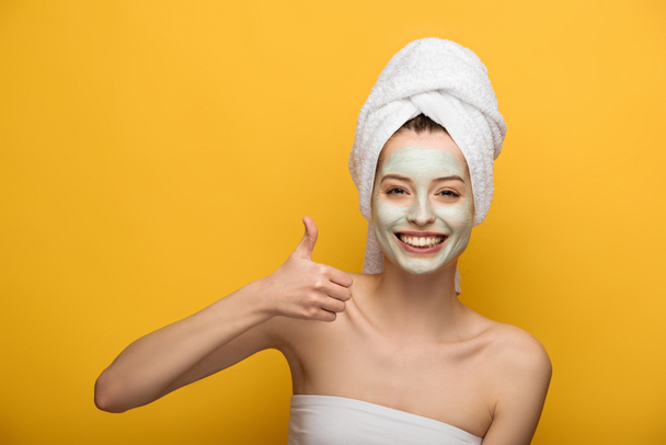 happy girl with nourishing facial mask showing thumb up and smiling at camera on yellow background - Photo, Image