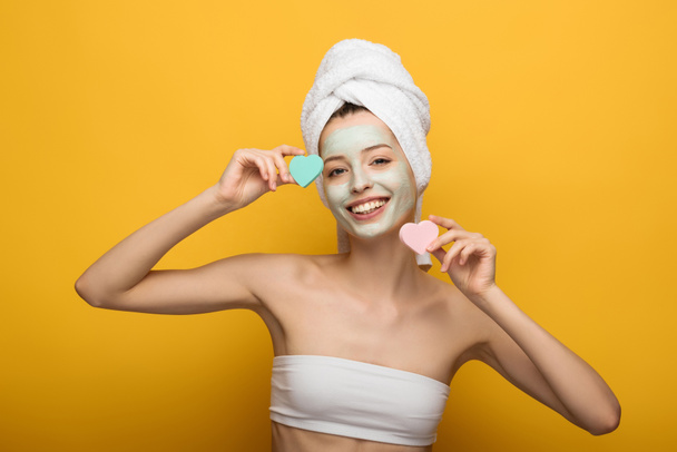 glückliches Mädchen mit nährender Gesichtsmaske mit herzförmigen Kosmetikschwämmen und Blick in die Kamera auf gelbem Hintergrund - Foto, Bild