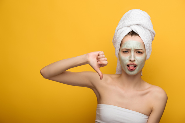 displeased girl with nourishing facial mask showing thumb down and sticking out tongue up on yellow background - Foto, imagen