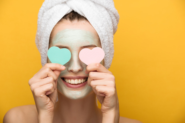 smiling girl with nourishing facial mask covering eyes with heart-shaped cosmetic sponges on yellow background  - Photo, Image