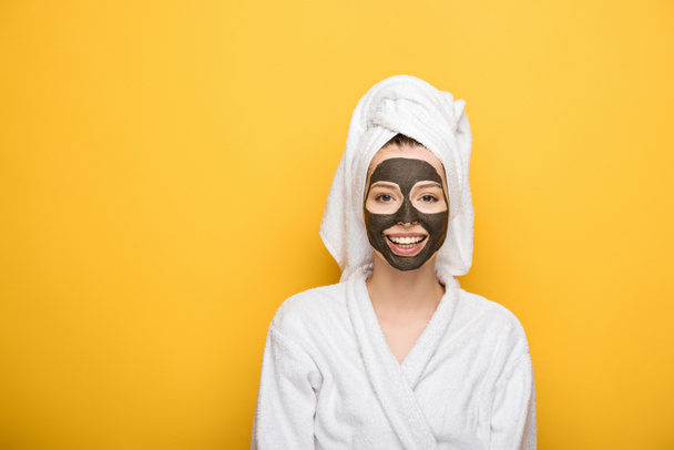 glimlachend meisje met gezicht klei masker en handdoek op hoofd kijken naar camera op gele achtergrond - Foto, afbeelding