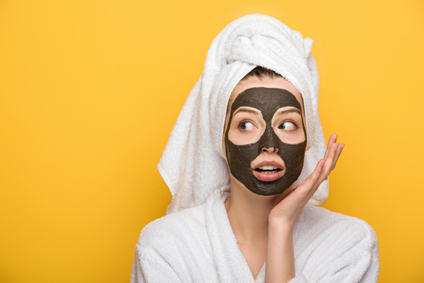 surprised girl with facial clay mask and towel on head holding hand near face isolated on yellow - Foto, Bild