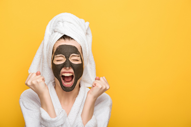 irritated girl with facial clay mask and towel on head holding clenched fists and screaming isolated on yellow - Photo, Image