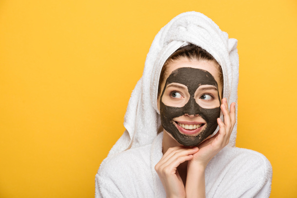 happy girl with facial clay mask and towel on head looking away isolated on yellow - Фото, изображение