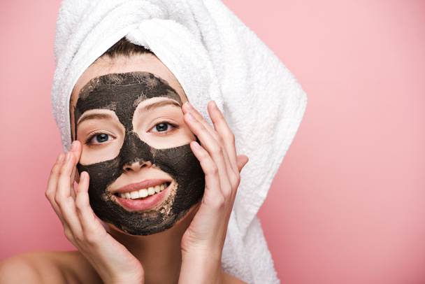 smiling girl with facial clay mask touching face and looking at camera isolated on pink - Photo, Image