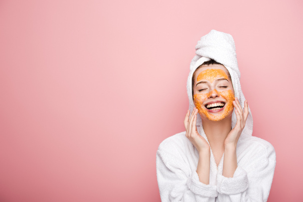 glückliches Mädchen mit Zitrusgesichtsmaske berührt Gesicht mit geschlossenen Augen isoliert auf rosa - Foto, Bild