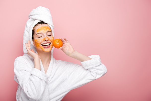 happy girl with citrus facial mask holding tangerine near face on pink background - Photo, image