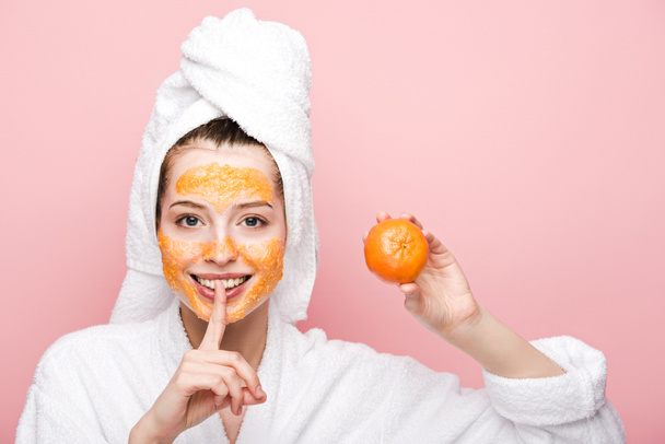positive girl with citrus facial mask holding tangerine and showing hush gesture isolated on pink - Foto, immagini