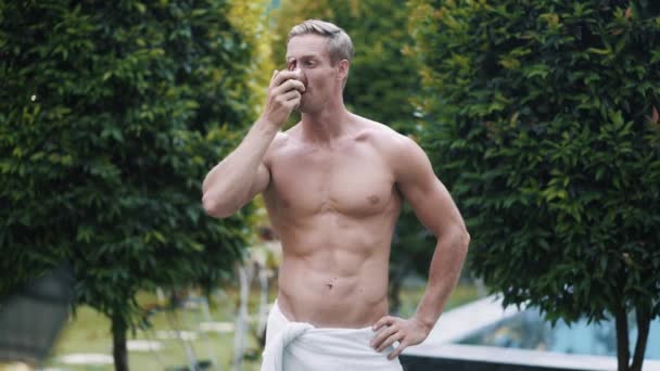 Portrait of sexy muscular shirtless man drinks coffee, greenery on background - Video