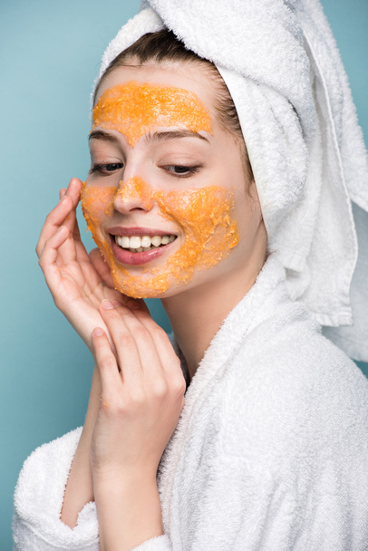 vrolijk meisje met citrus gezichtsmasker aanraken gezicht geïsoleerd op blauw - Foto, afbeelding
